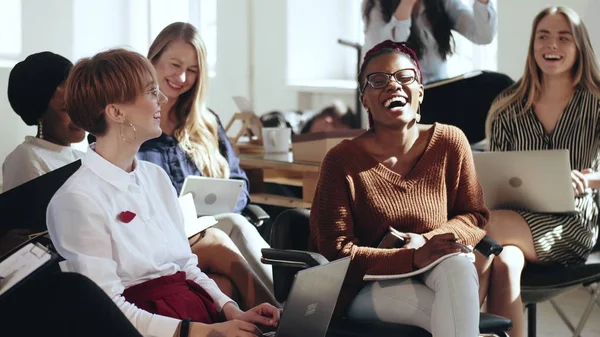 Felices jóvenes empresarias multiétnicas se sientan juntas escuchando el seminario corporativo riéndose en una conferencia de oficina moderna . — Foto de Stock