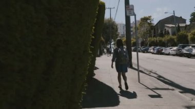 Kamera, yaz boyunca San Francisco caddesinde yeşil çitlere dokunarak gezen sırt çantalı mutlu genç turist kadını takip ediyor..