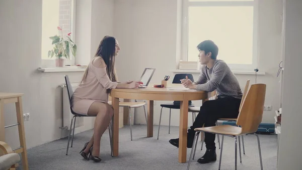 Young Asian man talking to confident businesswoman by the table. Multiethnic people talk in trendy modern office 4K.