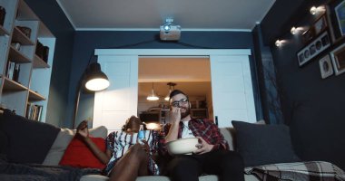 Mutlu, genç, çok ırklı romantik çift evde oturup patlamış mısırlı televizyon seyrediyor ve ağır çekimde gülüyorlar.