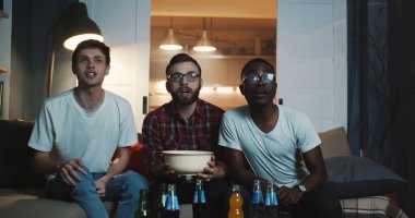 Mutlu çoklu etnik erkekler spor maçı izler, büyük projektör yavaş çekim kullanarak evde atıştırmalıklarla tezahürat yaparlar.