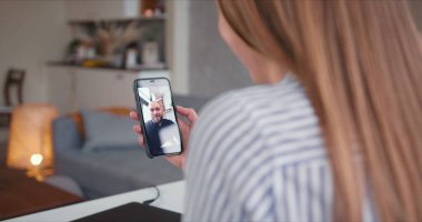 Omuz üstünden POV, akıllı telefon kullanan genç mutlu sarışın kadın evden bir arkadaşıyla görüntülü konuşma yapıyor..