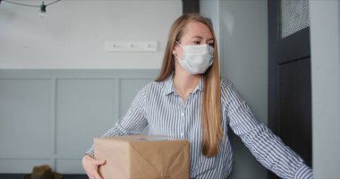 Tıbbi maskeli genç beyaz kadın virüs karantinası sırasında kuryeden paket teslimatı almak için daire kapısını açıyor..