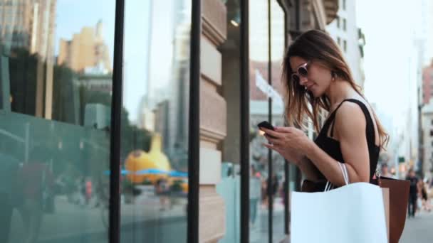 Jonge mooie vrouw met een zonnebril die tassen vasthoudt en met een smartphone naar de etalage kijkt. Langzame beweging. — Stockvideo