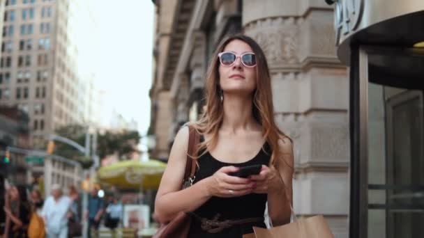 Retrato de una joven hermosa mujer usando un teléfono inteligente, sosteniendo bolsas de compras y caminando en el centro. Movimiento lento . — Vídeos de Stock