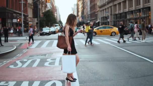 Jonge mooie vrouw met boodschappentassen die de straat oversteken in New York, USA en de smartphone gebruiken. Langzame beweging. — Stockvideo