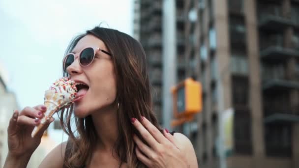 Portret młodej kobiety w okularach przeciwsłonecznych jedzącej topniejące lody z dodatkiem, delektującej się deserem w centrum. — Wideo stockowe