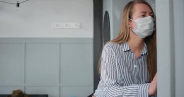 Tıbbi maskeli genç beyaz kadın virüs karantinası sırasında kuryeden paket teslimatı almak için daire kapısını açıyor..