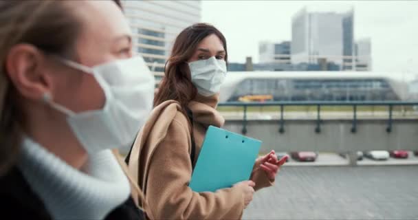 Vista lateral duas mulheres médicas caucasianas jovens usando máscaras faciais, andando na rua da cidade vazia com pranchetas em confinamento . — Vídeo de Stock