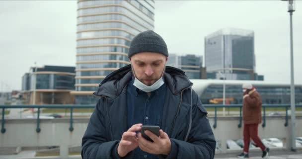 Эпидемический кризис. Портрет молодого бородатого белого мужчины с помощью смартфона, надевающего медицинскую маску на лицо на открытом воздухе — стоковое видео