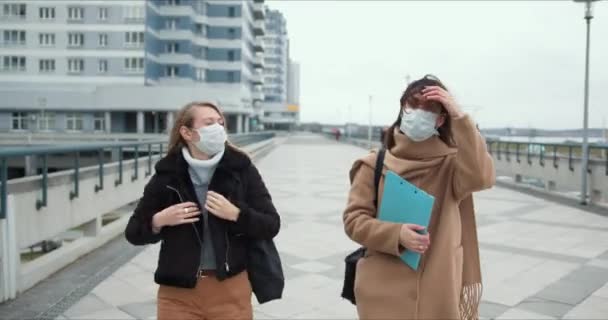 Dos mujeres médicas jóvenes usan máscaras caminando en la calle de la ciudad ventosa vacía. Atmósfera de miedo y peligro en cuarentena epidémica . — Vídeos de Stock