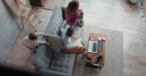 Top View dvě dospívající děti řídit jejich mladá matka blázen, zatímco ona se snaží pracovat z domova on-line pomocí notebooku. — Stock fotografie