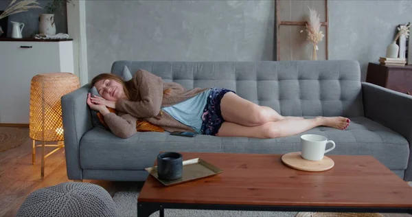 Glückliche junge schöne kaukasische blonde Frau, die ihr Smartphone zu Hause auf dem gemütlichen Sofa benutzt, sich hinlegt, um zu entspannen und ein Nickerchen zu machen. — Stockfoto