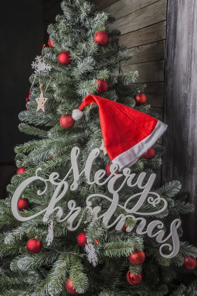 white merry christmas sign and red hat on decorated christmas tree on wooden background