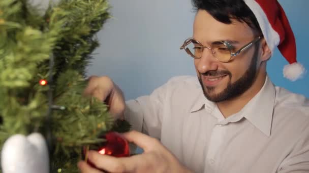 Lächelnder Junger Mann Mit Weihnachtsmann Hut Und Weihnachtsbaum Dekoration Drinnen — Stockvideo