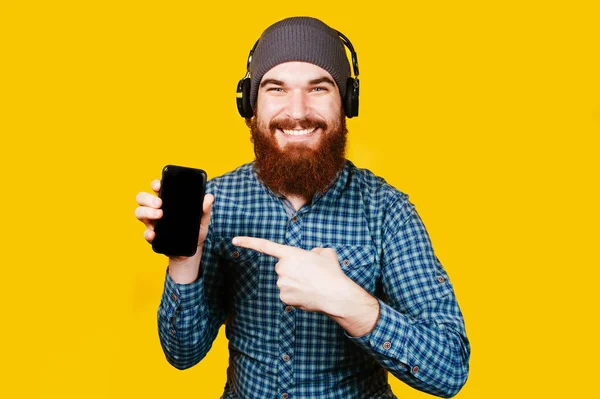 快乐的大胡子男子指着智能手机在橙色背景 用手拿手机的胡子微笑的人 演播室射击 带蓝牙耳机耳机的男士 — 图库照片