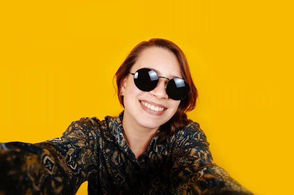 Cheerful happy young woman with sunglasses looking at camera and taking selfie on orange background in studio. Smiling happy girl.