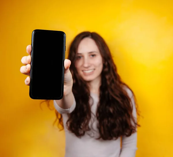 Mulher morena alegre tela do smartphone em branco e olhando para a câmera sobre o fundo amarelo. Foco seletivo . — Fotografia de Stock