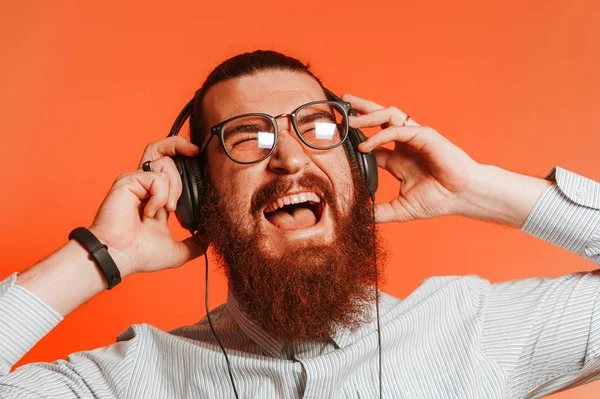 Porträtt av glada unga hipster skäggig man med hörlurar och glasögon på röd bakgrund. Ler man i avslappnad musik. — Stockfoto