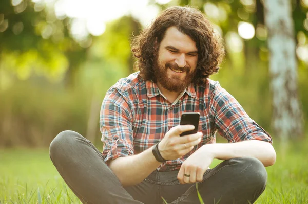 Alegre barbudo hipster hombre sentado fuera en el parque en la hierba y —  Fotos de Stock