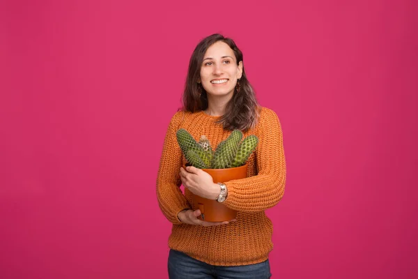 Retrato de una joven mujer feliz sosteniendo una planta de cactus grande mientras está de pie sobre el fondo rosado — Foto de Stock