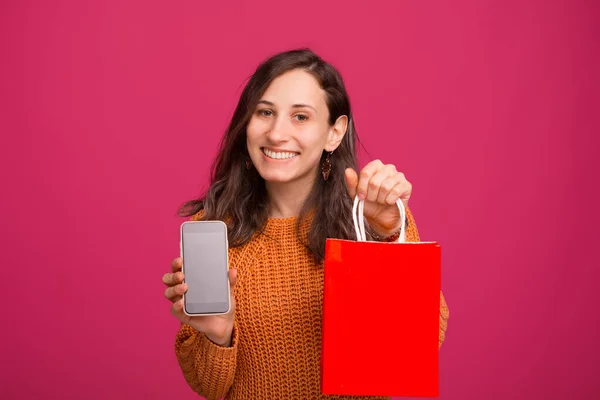 Glad ung kvinna som visar smartphone skärmen och hålla röd väska över rosa bakgrund — Stockfoto