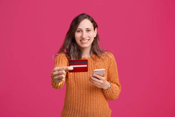 Menina encantadora em pé sobre fundo rosa e mostrando seu cartão de crédito e segurando smartphone — Fotografia de Stock