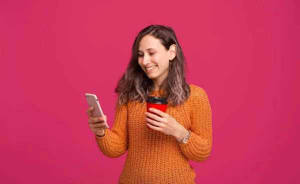 Joven mujer alegre de moda mirando su teléfono inteligente y sosteniendo la taza de café — Foto de Stock