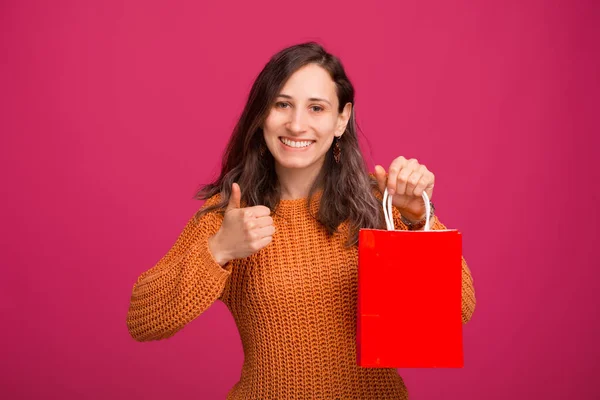 Jovem alegre vestindo suéter amarelo segurando saco de compras vermelho e mostrando polegares para cima — Fotografia de Stock
