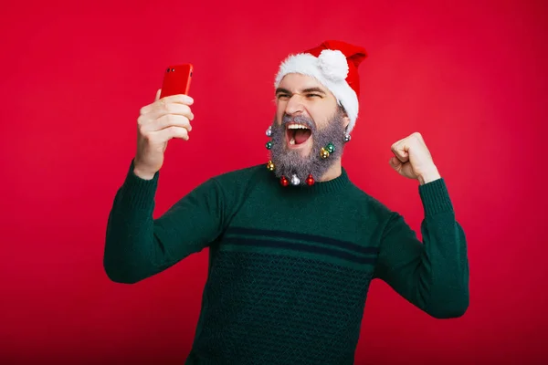 산타 클로스 모자를 쓰고 스마트폰을 보며 성공을 축하하는 수염을 기른 젊은이 — 스톡 사진