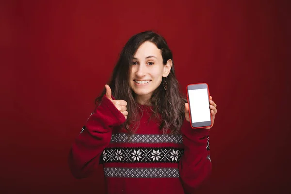 Портрет женщины в красном свитере, держащей смартфон с пустым экраном и показывающей большой палец на красном фоне — стоковое фото