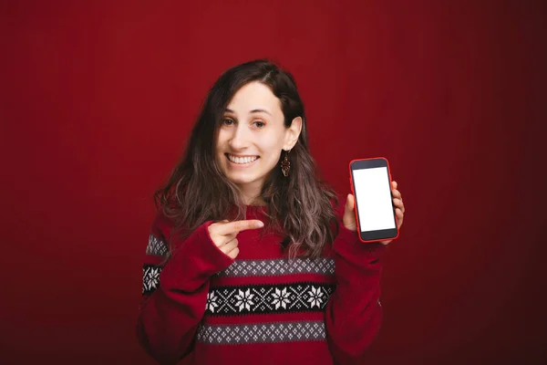 Vidám fiatal nő piros pulóvert visel és az okostelefon képernyőjére mutat — Stock Fotó