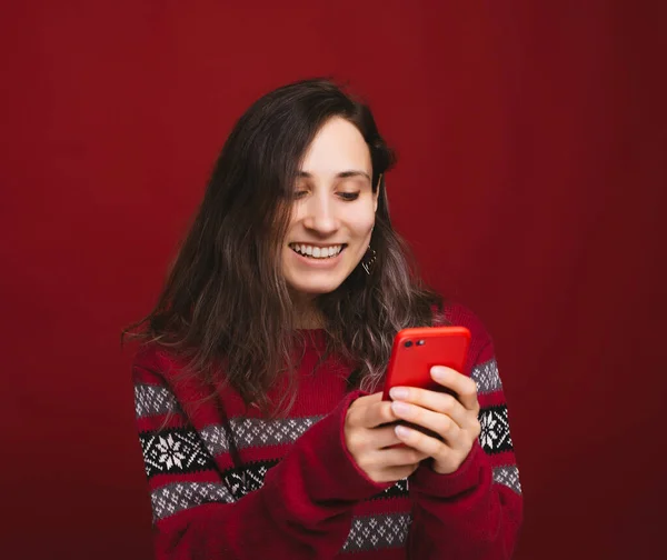 Mujer yougn alegre en suéter rojo usando su teléfono inteligente sobre fondo rojo — Foto de Stock