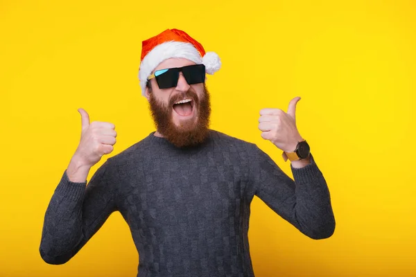 Wesoły przystojny brodaty hipster człowiek w kapeluszu Świętego Mikołaja pokazując kciuki na żółtym tle — Zdjęcie stockowe