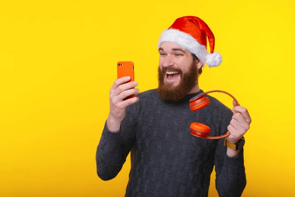 Ενθουσιασμένος άντρας με γενειάδα φορώντας το καπέλο του Αϊ Βασίλη και κοιτάζοντας smartphone και κρατώντας κόκκινα ασύρματα ακουστικά — Φωτογραφία Αρχείου