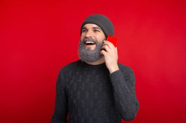 Mutlu sakallı hippi kırmızı arka planda duruyor ve akıllı telefondan konuşuyor.