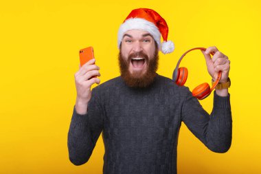 Noel Baba şapkası takan, elinde kırmızı akıllı telefon ve kablosuz kulaklık tutan harika sakallı adam.