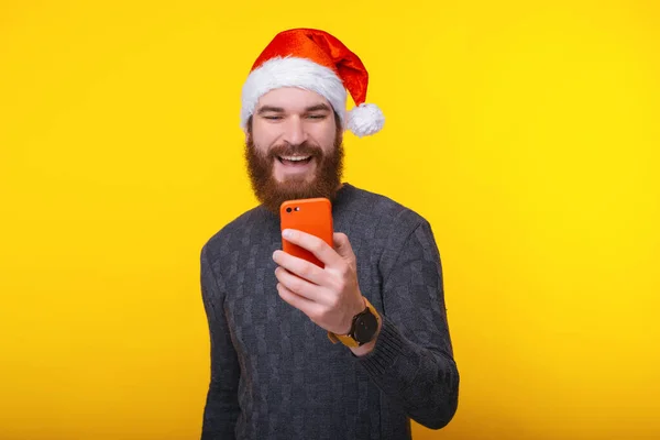 Souriant jeune homme barbu portant santa claus chapeau et en utilisant son nouveau téléphone mobile — Photo