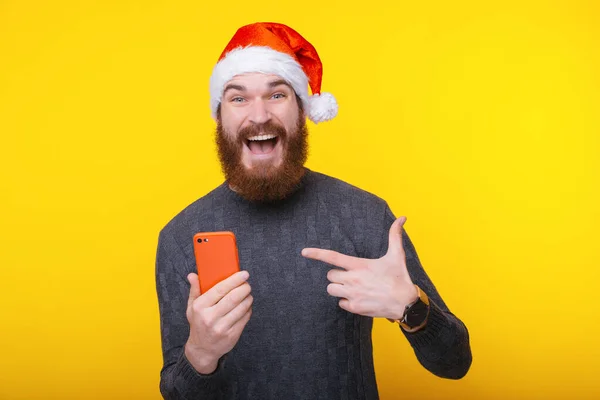 Amazed bearded hipster man in santa claus hat pointing red smartphone — ストック写真
