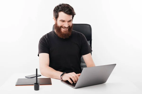 Wesoły uśmiechnięty brodaty mężczyzna siedzi w biurze i pracuje w la — Zdjęcie stockowe