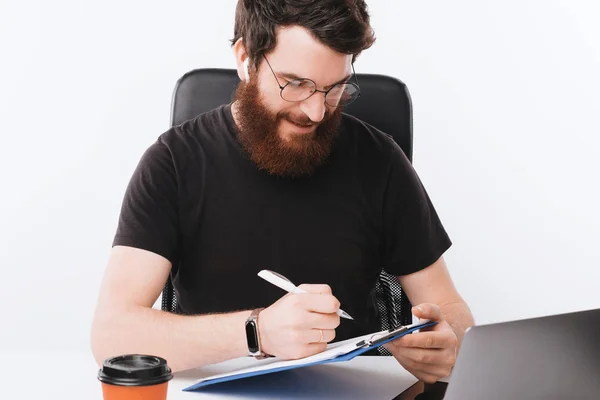 Foto de hombre feliz con barba sentado en la oficina y haciendo papel —  Fotos de Stock