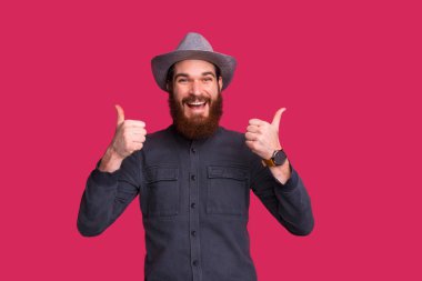 Mutlu sakallı adam, kafasında şapkayla kameraya baş parmağını gösteriyor.,