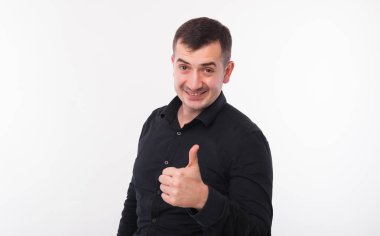Siyah gömlekli neşeli genç adam gülümsüyor ve beyaz arka planda baş parmağını gösteriyor..