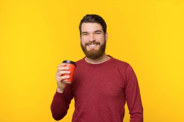 Sakallı adam gülümsüyor ve sarı arka planda bir fincan sıcak içecek tutuyor..