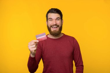 Kırmızı kazaklı sakallı mutlu adam sarı arka planda bir kredi kartı tutuyor..