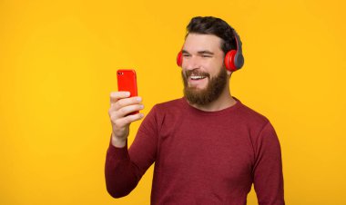 Sakallı genç adam müziği dinliyor, gülümsüyor ve sarı arka fonun yanındaki telefona bakıyor..