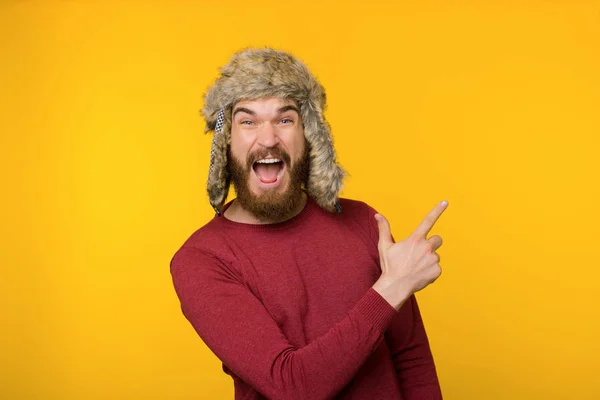 Brodaty facet w śmiesznej czapce zimowej, krzyczy i wskazuje w górę — Zdjęcie stockowe