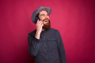 Genç sakallı adam başka tarafa bakıyor, gülümsüyor ve pembe duvarın yanında telefonla konuşuyor..