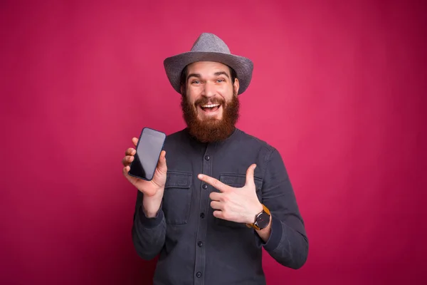 Jovem incrível está olhando para a câmera e mostrando um smartphone perto de uma parede rosa . — Fotografia de Stock