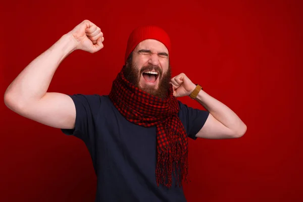 Brodaty, podekscytowany mężczyzna w szaliku i kapeluszu krzyczy z radości przy czerwonej ścianie.. — Zdjęcie stockowe